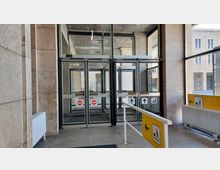 Eingangsbereich mit drehbaren Glastüren, die durch Schilder mit Richtungspfeilen und Durchfahrtsverboten markiert sind. Der Bereich ist von Steinwänden umgeben, mit Heizkörpern an der Seite und gelben Wegweisern mit Piktogrammen für Barrierefreiheit.