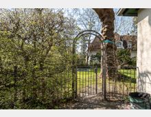 Metalltor mit Rundbogen eingerahmt von dichtem Buschwerk, das in einen Garten mit Rasen und Bäumen führt. Im Hintergrund ist ein großes Wohnhaus mit Satteldach und weißen Wänden zu sehen.