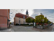 Straßenszene in einer Altstadt mit einem großen, beigefarbenen Gebäude mit rotem Satteldach und Kirchturm im Hintergrund. Im Vordergrund stehen Bäume in einer gepflasterten Platzanlage mit einer Tourist-Informations-Tafel.