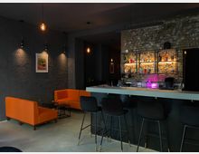 Barbereich mit moderner Einrichtung: Eine schwarze Theke mit hohen Barhockern, eine beleuchtete Backsteinwand mit Regalen voller bunter Flaschen, daneben orangefarbene Sofas und kleine Couchtische. Dezente Beleuchtung und eine dunkelgraue Wand mit einem gerahmten Bild sorgen für eine stimmungsvolle Atmosphäre.