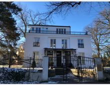 Weiße Villa mit drei Stockwerken und Flachdach, umgeben von kahlen Bäumen und einem schmiedeeisernen Zaun. Der Vorgarten ist teilweise mit Schnee bedeckt, und ein gepflasterter Weg führt zum Eingang des Hauses.