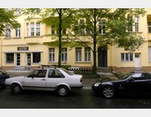 Gelbes Wohngebäude mit großen Fenstern und einem Geschäft mit der Aufschrift „Antik“ im Erdgeschoss. Vor dem Gebäude stehen parkende Autos und Bäume säumen den Gehweg.