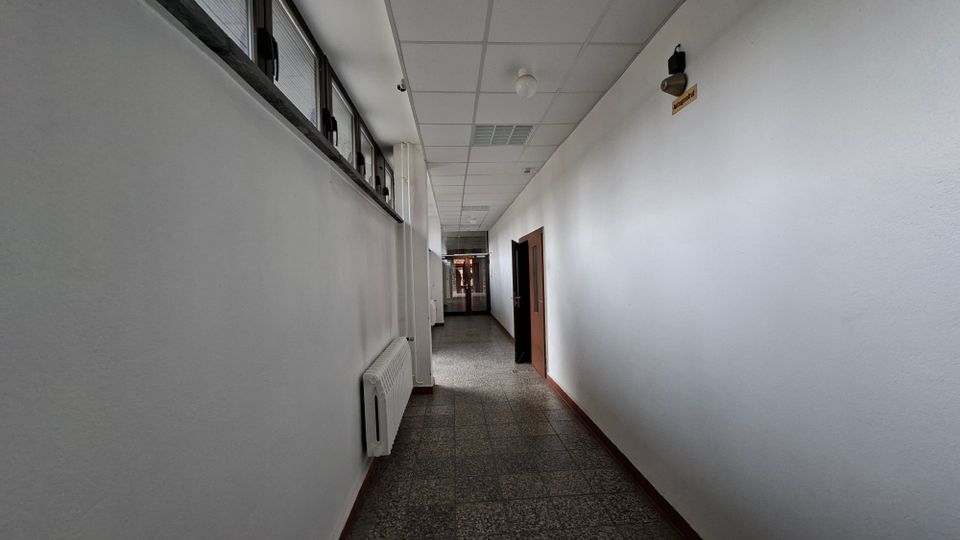 Langer, schmaler Innenflur in einem Gebäude mit weißen Wänden, grauem Steinboden und abgehängter Rasterdecke mit Leuchten. Links befinden sich hohe Fenster und ein Heizkörper, rechts mehrere Türen; am Ende führt eine verglaste Tür in einen weiteren Bereich.