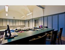 Innenaufnahme eines Sitzungssaals mit grüner Holzvertäfelung und einer gewölbten Decke. Im Vordergrund ein großer Tisch mit Computer, Mikrofon und Schreibutensilien, umgeben von Holzstühlen; im Hintergrund sind Türen und eine Uhr an der Wand sichtbar.