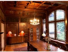 Ein elegantes Esszimmer mit reich verzierten Holzvertäfelungen und einem kunstvollen Kassettendeckenmuster. Eine große Fensterfront mit farbigen Glasdetails lässt Tageslicht in den Raum, der mit einem Holztisch, Stühlen und dekorativen Lampen ausgestattet ist.