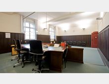 Ein Gerichtssaal mit dunklen Holzvertäfelungen an den Wänden, einem großen Holztisch mit mehreren Stühlen und Akten darauf. Im Hintergrund befinden sich Reihen von Stühlen, eine rote Tür und hohe Fenster, die natürliches Licht einlassen.