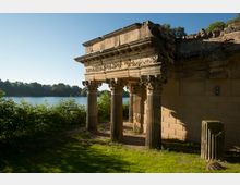 Ruinenartiges Bauwerk mit steinernen Säulen und verzierten Kapitellen, das an eine antike Tempelarchitektur erinnert, gelegen in einer grasbewachsenen Umgebung am Ufer eines Sees, im Hintergrund von Bäumen umgeben.