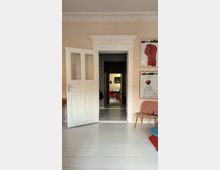 Interior of a home with pale walls, white-painted wood floors, and an ornate white doorway with decorative molding and a partially open double door leading to a darker hallway and another room beyond. On the right wall are two framed posters, and a low chair sits near the corner.
