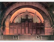 Innenraum einer Kirche mit einer großen Orgelanlage vor einer halbrunden Steinwand. Die Orgel ist verziert mit detaillierten Holzschnitzereien und zahlreichen Pfeifen, beleuchtet von warmem Licht.