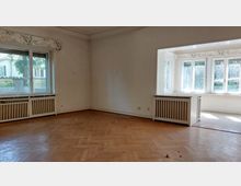 Ein leerer Raum mit Parkettboden und großen Fenstern. Links ein einfaches Fenster mit Heizkörperverkleidung, rechts ein Erker mit mehreren Fenstern, die viel Tageslicht hereinlassen.