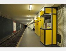 U-Bahn-Station mit einem langen Bahnsteig, Schienen auf der linken Seite und gelben Wandpaneelen rechts, die eine kleine Kabine umschließen. Der Bereich ist beleuchtet, mit mehreren Sitzbänken und vereinzelten Personen entlang der weißen Ziegelwand rechts.