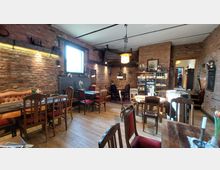 Gemütliches Café oder Restaurant mit Backsteinwänden, Holzboden und einer Mischung aus gepolsterten Sitzbänken und Holzstühlen. Im Raum befinden sich verschiedene Tische, eine Regalwand mit Verkaufsartikeln sowie ein Fenster, das Tageslicht hineinlässt.