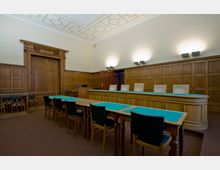 Gerichtssaal mit holzgetäfelten Wänden, einer Richterbank mit vier Stühlen und Tischen mit grünem Einlagebezug im Vordergrund. Der Raum verfügt über eine dekorative Tür mit Holzornamentik und eine helle Decke mit geometrischen Mustern.