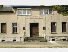 Fassade eines schlichten, rechteckigen Gebäudes aus der Mitte des 20. Jahrhunderts mit einer symmetrischen Gestaltung. Der Eingang wird durch eine Treppe mit Geländern und gemusterten Steinverzierungen eingefasst, darüber steht die Inschrift „POS. Karl Liebknecht 1955“.