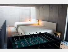 Minimalistisches Schlafzimmer mit Sichtbetonwänden und Holzfußboden, zwei großen Betten mit weißen Decken und schlichten Kopfteilen sowie warmen Wandlampen. Im Vordergrund steht ein Tischkicker, daneben ein kleiner Hocker nahe einem Fenster.