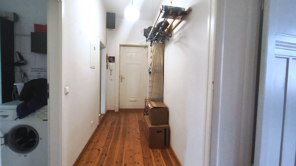 Narrow apartment hallway with white walls and a wooden plank floor leading to a white door at the end, lit by a hanging ceiling lamp. A wall-mounted rack with hangers and a few cardboard boxes lines the right side, and an open doorway on the left reveals a small laundry area with a washing machine.