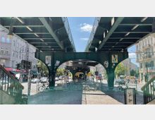 Ansicht einer belebten Straßenszene in einer Stadt mit einer grün lackierten Stahlkonstruktion, die eine erhöhte Bahnstrecke trägt. Unter der Konstruktion befindet sich eine Straße mit geparkten Fahrrädern, mehreren Autos und Fußgängern, eingerahmt von Gebäuden mit Geschäften und Wohnungen.