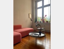 Wohnzimmer mit Holzboden, einem rosa Sofa, einem runden Couchtisch mit einer Vase und einem Tablett darauf. Im Hintergrund ein großes Fenster mit Zimmerpflanzen auf der Fensterbank und ein Hocker mit einer Decke daneben.