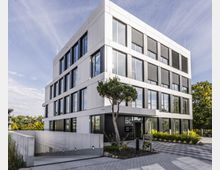 Modernes Bürogebäude mit vier Stockwerken, großen Fenstern und weißen Fassadenelementen. Vor dem Gebäude befinden sich ein kleiner Baum, gepflasterte Wege und eine Einfahrt zur Tiefgarage.