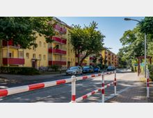 Mehrstöckige Wohngebäude mit gelben Fassaden und roten Balkonen in einer von Bäumen gesäumten Straße. Im Vordergrund geparkte Autos, ein Gehweg, eine Absperrung mit roten und weißen Streifen sowie eine Bushaltestelle.