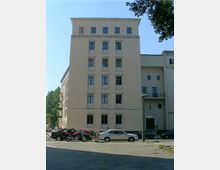 Mehrstöckiges Wohngebäude mit heller Fassade und symmetrisch angeordneten Fenstern, gelegen an einer Straße mit geparkten Autos und Motorrädern. Im Vordergrund ein Gehweg und ein beleuchteter Straßenbereich; Bäume sind am Bildrand sichtbar.