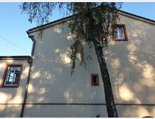 Zweigeschossiges Gebäude mit beigefarbener Fassade und rot-braunen Fensterrahmen, vor dem ein großer Baum steht. Sonnenlicht wirft Schatten der Äste auf die Wand.