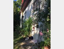 Ein weiß gestrichenes Wohngebäude mit großen Fenstern, umgeben von Rankrosen und grünen Pflanzen. Im Vordergrund stehen mehrere Blumentöpfe, eine Laterne und ein Gartenrechen auf einem mit Steinplatten ausgelegten Weg; dahinter erstreckt sich ein gepflegter Garten.