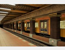 Eine U-Bahn-Station mit Fliesenboden und dunkelroten Säulen, die den Bahnsteig säumen. Ein gelber U-Bahn-Wagen fährt durch die Station, während die Decke mit linearen Leuchtstofflampen beleuchtet ist.