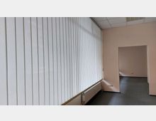 Innenraum eines leeren Büros mit einer großen Fensterfront, die von vertikalen Jalousien bedeckt ist. Der Raum hat beige Wände, einen grauen Boden, eine weiße Heizung unter der Fensterfront und ein angrenzendes, ebenfalls leeres Zimmer sichtbar durch eine Türöffnung.