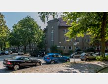 Wohnstraße mit mehrstöckigem Wohngebäude im Hintergrund, das eine beige Fassade und ein rotes Ziegeldach hat. Im Vordergrund stehen mehrere geparkte Autos auf einer gepflasterten Straße, umgeben von großen Bäumen, die Schatten spenden.