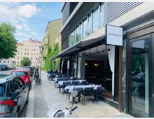 Straßenszene mit einem Restaurant mit offener Glasfront und Außenbestuhlung, beschriftet mit "Osterberger Mitte". Links sind parkende Autos und rechts mehrstöckige Gebäude, teils begrünt.