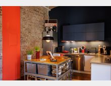 Moderne Küche mit einer Kombination aus freiliegender Ziegelwand und dunkelblauen Schränken. Eine zentrale Kücheninsel mit offenem Regal, eine Edelstahl-Arbeitsplatte, ein Hängelicht und verschiedene Küchenutensilien sowie Pflanzen ergänzen das Design.