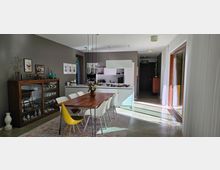 Modernes Esszimmer mit angrenzender Küchenzeile, bestehend aus einem Holztisch, umgeben von weißen und einem gelben Stuhl, sowie einer Vitrine mit Geschirr an der grauen Wand. Rechts befinden sich große Glas-Schiebetüren mit Vorhängen, die Tageslicht in den Raum lassen.