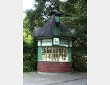 Ein kleiner Pavillon mit spitz zulaufendem Dach, grünem Holzrahmen und roten Kacheln an der Fassade, umgeben von Bäumen. Im Schaufenster sind Keramikartikel ausgestellt, über dem Fenster befinden sich Schilder mit der Aufschrift „Keramik“ und „Sabine Martin“.