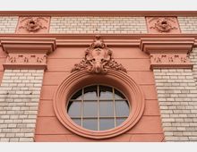 Nahaufnahme einer historischen Backsteinfassade mit rosafarbenen Stuckelementen und einem runden Fenster mit Sprossen. Über dem Fenster befindet sich ein reich verziertes Ornament mit Maskenrelief und floralen Details.