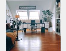 Modernes Arbeitszimmer mit Schreibtisch vor zwei Dachfenstern, einem schwarzen Bürostuhl und weißen Regalen an der rechten Wand. Der Raum ist hell mit Holzboden, großen Zimmerpflanzen und dekorativen Elementen wie einer Stehlampe und Bildern.