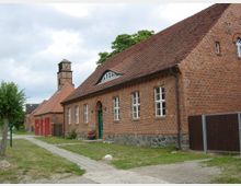 Historisches Backsteingebäude mit rotem Ziegeldach, mehreren rechteckigen Fenstern und einem kleinen Glockenturm im Hintergrund. Vor dem Gebäude verläuft ein gepflasterter Weg, umgeben von Rasenflächen und Bäumen.