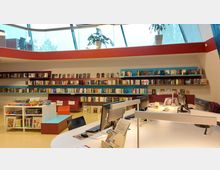Innenraum einer modernen Bibliothek mit bunten Bücherregalen an der Wand, einem großen Tisch mit mehreren Computerarbeitsplätzen und einem kleinen Bereich mit Brettspielen. Über dem Raum befinden sich große Fenster mit schrägen Glaselementen und Pflanzen.