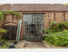 Backstein-Gebäude mit Satteldach, einer großen Glastür an der rechten Seite und einem alten Holztor links. Efeu und andere Pflanzen wachsen entlang der Fassade und um das Gebäude herum.