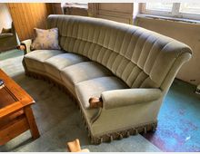 Gebogener Wohnraum mit großem, gepolstertem Samtsofa in hellem Grau mit Fransenbesatz und Holzarmlehnen, davor ein Holztisch. Im Hintergrund stehen hohe Fenster und eine holzvertäfelte Wand, der Boden ist teils mit Teppich, teils mit abgenutztem Anstrich versehen.