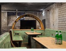 Industrie-inspirierter Innenraum mit Sitzbänken aus verwittertem grünem Holz, rustikalen Holztischen und einer großen, gelblich rostigen Metallbogenstruktur im Hintergrund. Umgeben von Backstein- und Betonwänden, ergänzt durch moderne Elemente wie einen Flachbildschirm und Wasserflaschen samt Gläsern auf dem Tisch.