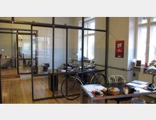 Ein modernes Büro mit großen Fenstern, das durch Metall-Glas-Trennwände in Arbeitsbereiche unterteilt ist. Im Vordergrund steht ein Schreibtisch mit Arbeitsmaterialien, im Hintergrund sind ein Fahrrad, weitere Schreibtische und Büromaterialien erkennbar.