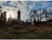 Eine ländliche Szene mit einem hohen Backsteinturm mit Kuppeldach im Hintergrund, umgeben von überwucherten Pflanzen und kahlen Bäumen. Im Vordergrund befindet sich eine Wiese mit einem Pfad, während im Hintergrund vereinzelte Gebäude sichtbar sind.