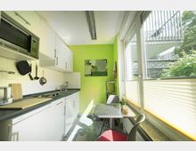 Kleine moderne Küche mit weißer Einbauküche, grün gestrichener Wand und einem kleinen Esstisch vor einem großen Fenster mit Blick ins Grüne. An der Wand hängen Küchenutensilien, und durch das Fenster fällt Tageslicht in den Raum.