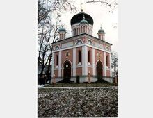 Ein kleines orthodoxes Kirchengebäude mit rosa-weißer Fassade, drei Kuppeln mit goldenen Kreuzen und Rundbogenfenstern. Die Kirche steht in einer winterlichen Landschaft mit kahlen Bäumen und schneebedecktem Boden.