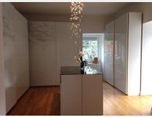 Minimalistischer Raum mit glänzend weißen Einbauschränken an den Wänden, einer Kücheninsel mit dunkler Arbeitsfläche in der Mitte und einem Holzfußboden. Im Hintergrund ist ein Fenster mit Blick ins Freie, dekoriert mit Blumen und kleinen Gegenständen.