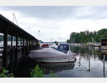 Ein kleiner Hafen mit überdachten Anlegestegen, mehreren abgedeckten Booten und ruhig liegendem Wasser. Im Hintergrund sind weitere Boote, Bäume und bewaldete Ufer zu sehen, unter einem bewölkten Himmel.
