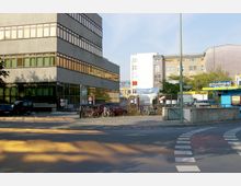 Mehrstöckiges Bürogebäude aus grauem Beton mit großen Fensterflächen, das an einer Kreuzung liegt. Im Vordergrund sind parkende Fahrräder, Autos und ein blauer Kiosk sichtbar, im Hintergrund stehen weitere mehrstöckige Wohn- und Geschäftshäuser.