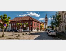 Straßenszene in einer Altstadt mit Kopfsteinpflaster, flankiert von historischen Gebäuden, darunter ein rotes Haus im Vordergrund. Im Hintergrund ist der Turm einer Backsteinkirche zu sehen, während geparkte Autos und Bäume die Straße säumen.
