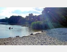 Ein kleiner Sandstrand an einem See mit klarem Wasser, in dem zwei Personen baden. Im Hintergrund stehen dichte Bäume und Schilf am Ufer, während die Sonne durch die Wolken strahlt.